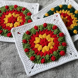 May include: Three handmade crochet coasters. Each coaster has a square white border with a colourful floral design in the centre. The floral design includes red, yellow, and green elements, creating a vibrant and textured appearance.