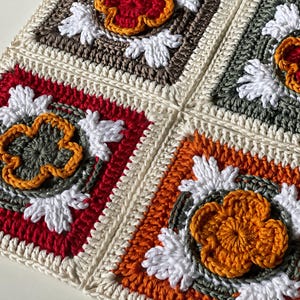 May include: Close-up of a crocheted blanket with four square panels. Each square features a central flower design in shades of red, orange, and yellow, surrounded by white and gray accents. The squares are bordered by cream-colored yarn.