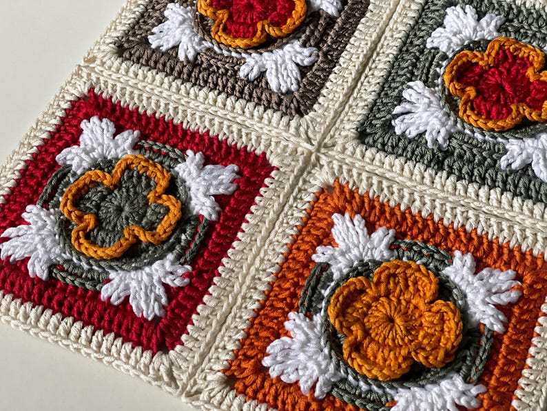 May include: Close-up of a crocheted blanket with four square panels. Each square features a central flower design in shades of red, orange, and yellow, surrounded by white and gray accents. The squares are bordered by cream-colored yarn.