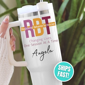 May include: White tumbler with a handle, featuring the text "RBT Registered Behavior Technician" in purple and orange. Below, it reads "Changing Lives One Session At A Time" and the name "Angela" in script.