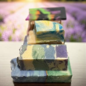 May include: A stack of handmade soaps with various colors and patterns. The soaps include shades of blue, green, yellow, purple, and black. The background features a blurred view of a lavender field and a sunset.