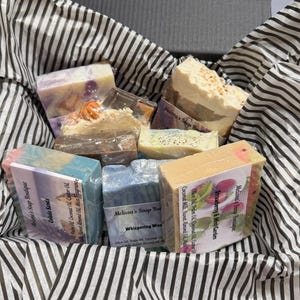 May include: A gift box filled with various handmade soaps. The soaps are in different colors and shapes, some wrapped with labels. The box is lined with black and white striped tissue paper, creating a visually appealing presentation.