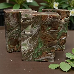 May include: Four rectangular bars of handmade soap with a marbled design. The soap features swirls of brown, green, and white, creating a natural, earthy aesthetic. The bars are arranged on a brown surface, with greenery and white flowers in the background.