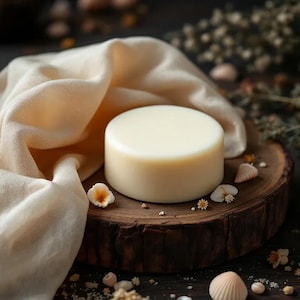 May include: A round, off-white bar of soap sits on a rustic wooden slice, surrounded by small seashells, dried flowers, and a cream-colored fabric. The soap has a smooth surface and a cylindrical shape, suggesting a natural skincare product.