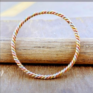 May include: A twisted metal bangle bracelet with a multicolored design. The bracelet features a spiral pattern with alternating sections of gold, silver, and copper-toned metal. The bracelet is displayed on a wooden surface.