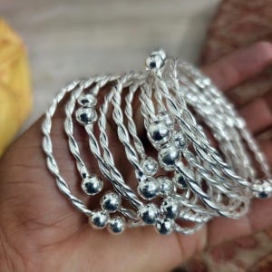 May include: A set of multiple silver-toned bangles featuring a twisted wire design. Each bangle is finished with two small, smooth, round silver-toned spheres at the open ends. The bracelets are stacked together, showcasing a reflective metallic surface.