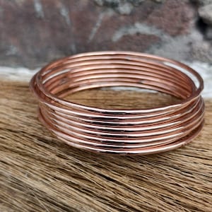 Könnte beinhalten: Ein Stapel kupferfarbener Armreifen. Die Armbänder sind rund und scheinen aus dünnem Metall zu bestehen. Die Armreifen sind übereinander gestapelt. Der Hintergrund ist eine Mischung aus Braun und Grau.