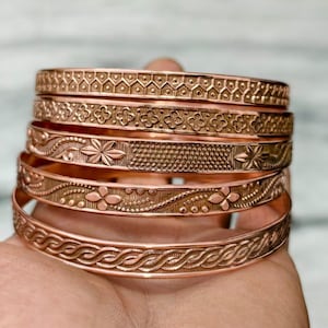 Può includere: Una pila di cinque braccialetti rigidi in metallo color rame, ognuno caratterizzato da un distinto motivo in rilievo. I design includono zigzag geometrici, motivi floreali a quattro petali, fasce punteggiate e un motivo a corda intrecciata.