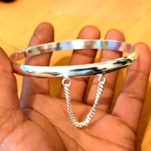 May include: A silver bangle bracelet with a safety chain. The bracelet is round and has a polished finish. The chain is made of small, twisted links. The bracelet is held in a hand.
