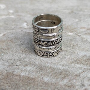 May include: A stack of three silver rings featuring intricate embossed floral and bird patterns. The interior of the top ring is clearly stamped with the hallmark 925, indicating sterling silver composition. Each band has a polished finish.