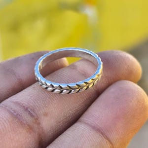 Peut inclure: Une bague en argent avec un motif de feuilles. La bague est tenue dans une main, avec un fond jaune flou. La bande de la bague présente un motif de feuilles répétitif.