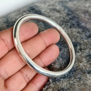 May include: A shiny, silver-colored bangle bracelet. The smooth, rounded bracelet is oval-shaped and appears to be made of metal. The bracelet is held in a hand, with a soft, textured background.