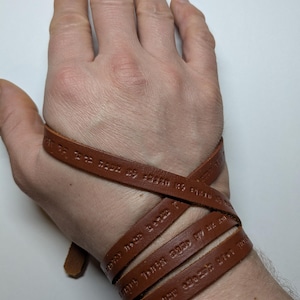 Peut inclure: Un bracelet en cuir marron à plusieurs brins, chacun avec une inscription différente. Le bracelet est enroulé autour du poignet d'une personne.