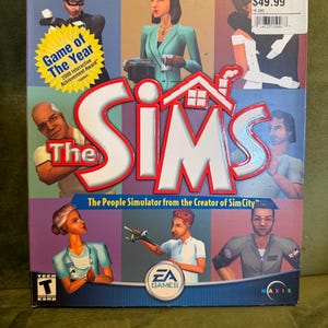 May include: The Sims video game box. The box features character illustrations and the game's logo in red and white. The box also includes the text "Game of The Year" and "The People Simulator from the Creator of SimCity."