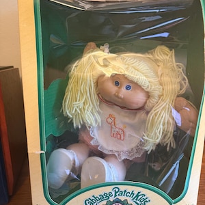 May include: A Cabbage Patch Kids doll in its original box. The doll has blonde yarn hair, blue eyes, and a bib with a giraffe design. The box is tan with green accents and the text "Cabbage Patch Kids".