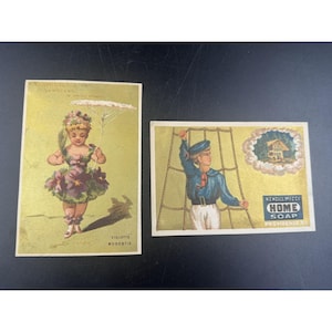 May include: Two vintage advertising cards. The first card features a young girl in a purple dress holding a parasol. The text on the card reads "Violette Modestie". The second card features a young boy in a sailor outfit climbing a rope ladder. The text on the card reads "Kendall Meeks Home Soap Providence R.I."