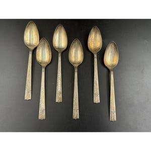 May include: A set of six silver-toned metal spoons arranged in two rows. Each spoon features an oval bowl and a slender handle with a decorative embossed pattern at the base. The metal has a slightly tarnished, aged finish with visible patina.