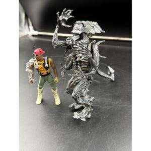 May include: Two action figures: a silver alien-like creature and a human figure in a red cap and vest. The human figure is wearing a vest with the words "NO BUGS". The alien figure has a detailed, textured design.