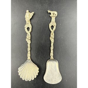 May include: Two silver-toned metal spoons with ornate, textured handles featuring floral motifs. The left spoon has a shell-shaped bowl and a figure at the top, while the right spoon has a bell-shaped bowl and a small animal figure at the top.