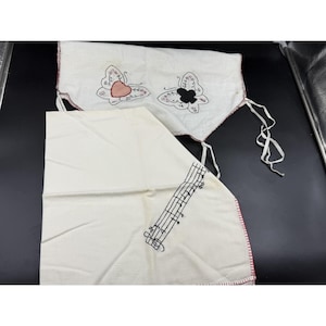 May include: Two white fabric items with red trim. One has two embroidered butterflies, one pink and one black. The other has embroidered musical notes. Both items have white ties.