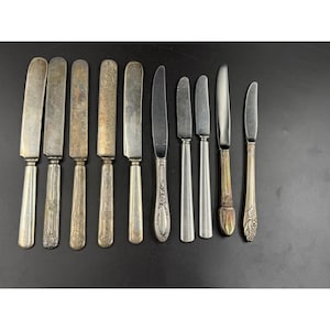 May include: A collection of antique butter knives with various handle designs and blade shapes. The knives are made of silver-toned metal and are arranged on a black surface. Some handles have ornate patterns.