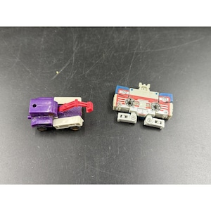 May include: Two vintage toy robots. One is a purple and white truck with a red crane arm. The other is a red, white, and blue robot with a cassette tape design. Both are on a dark surface.