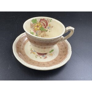 May include: Cream-colored teacup and saucer set with a floral design. The cup features a brown band around the middle and a curved handle. The saucer has a matching brown border with a scalloped edge. The set is likely vintage.