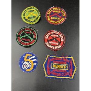 May include: A collection of vintage National Rifle Association (NRA) patches. The patches feature text such as "Marksman," "Sharpshooter," and "Junior." Colors include red, blue, yellow, green, and white. Some patches have crossed rifles.