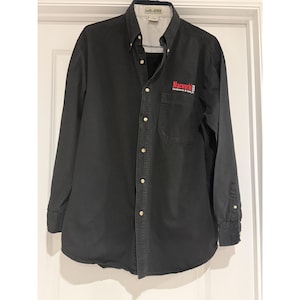 May include: A black long-sleeve button-down shirt featuring a collar, a front chest pocket, and light-colored buttons. The text Macworld Conference & Expo is embroidered in red and white on the chest pocket area of the garment.