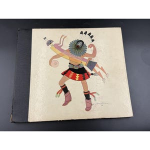 May include: A vintage book with a black spine and a cover featuring a colorful illustration of a Native American figure. The figure is adorned with a headdress, skirt, and boots, and is holding symbolic objects. The artwork is signed by Gerald Nailor.