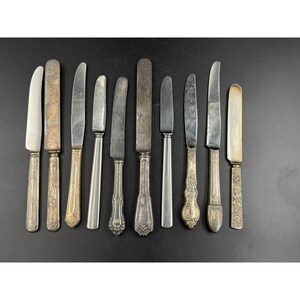 May include: A collection of antique butter knives with various handle designs. The blades are silver-toned, and the handles feature ornate patterns and textures. The knives are arranged on a dark surface, showcasing their vintage aesthetic.
