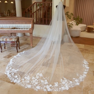 以下が含まれることがあります： 白い花のレーストリムが付いた、長く流れるような白いウェディングベール。マネキンに飾られたベールは、繊細で透き通った生地と、縁に沿った白い花のアップリケの複雑なディテールを際立たせています。クラシックなウェディングアクセサリーです。