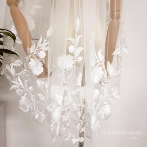 Puede incluir: Velo de novia blanco con delicado encaje floral. El encaje presenta un patrón repetitivo de flores y hojas blancas.