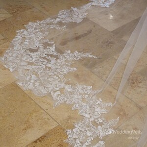 Peut inclure: Un voile de mariée blanc avec de délicates applications de dentelle florale. Le voile est fait de tulle doux et a un bord festonné. Les applications de dentelle sont dispersées sur tout le voile, créant un look romantique et élégant.