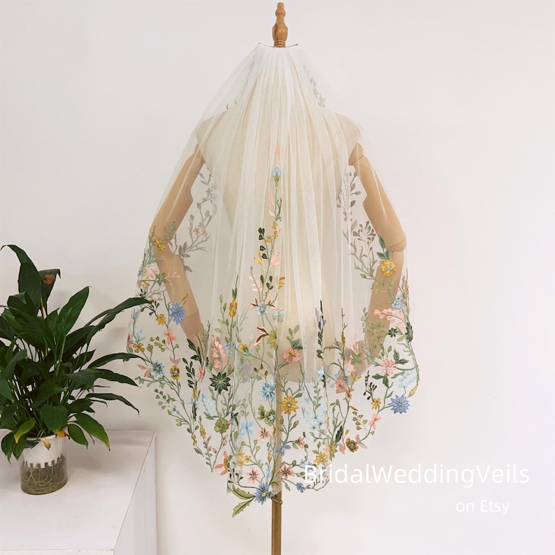 Pode incluir: Um v&eacute;u de noiva branco com um delicado bordado floral em tons de azul, verde, amarelo e rosa. O v&eacute;u &eacute; apresentado em um manequim.