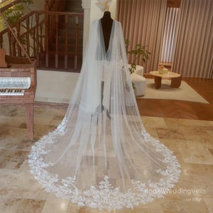 May include: A white tulle wedding veil with a delicate lace trim. The veil is long and flowing, with a deep V-neckline. The veil is displayed on a mannequin in a room with a piano and a staircase.