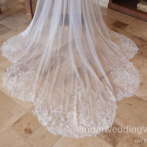 Peut inclure: Un long voile de mariée blanc et fluide avec des détails de dentelle florale complexes le long de l'ourlet. Le voile est fait d'un tissu transparent et léger, créant un look délicat et élégant. Le voile est présenté sur un sol carrelé.