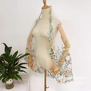 Pode incluir: Um v&eacute;u de noiva de tule branco com bordado floral delicado em tons de azul, amarelo e rosa. O v&eacute;u est&aacute; drapeado sobre um manequim e apresenta um design floral que se estende pelos lados.