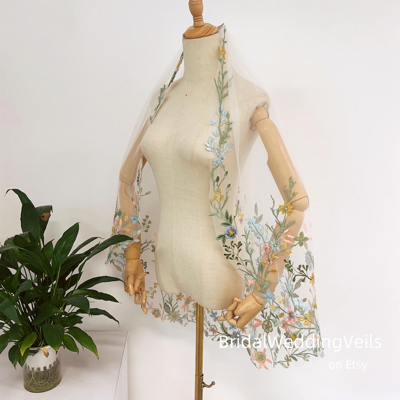 Pode incluir: Um v&eacute;u de noiva de tule branco com bordado floral delicado em tons de azul, amarelo e rosa. O v&eacute;u est&aacute; drapeado sobre um manequim e apresenta um design floral que se estende pelos lados.