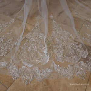 May include: A sheer white wedding veil with intricate, ornate lace detailing. The lace features floral and scroll patterns, embellished with sparkling sequins. The veil is spread out on a light-colored surface, showcasing the delicate design.