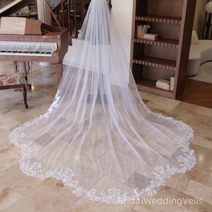 Peut inclure: Un long voile de mariée blanc avec une délicate bordure en dentelle florale sur le bord. Le voile est présenté sur un mannequin, mettant en valeur sa longueur et son design complexe. Le voile est fait d'un tissu transparent et fluide.
