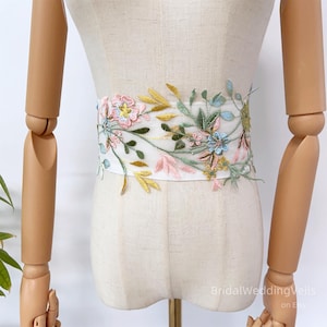 Könnte beinhalten: Ein weißer Brautschärpe mit zarter floraler Stickerei. Das gestickte Design zeigt rosa, blaue und gelbe Blumen mit grünen Blättern. Die Schärpe wird auf einer Schaufensterpuppe präsentiert.