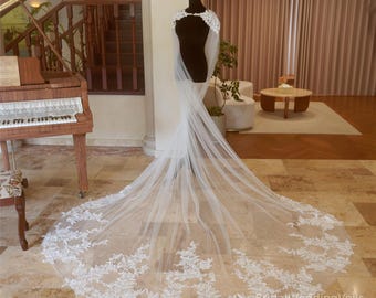 May include: A white tulle wedding veil with a lace trim. The veil is long and flowing, and it is draped over a mannequin. The veil is decorated with delicate floral lace.