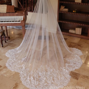 May include: A long, sheer white bridal veil with a delicate, embroidered lace border. The veil is displayed on a mannequin, showcasing its length and intricate design. The lace features a floral pattern with small, beaded accents.