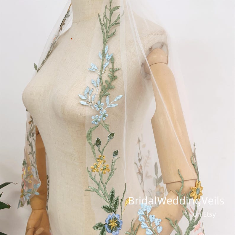 Pode incluir: Um v&eacute;u de noiva de tule branco com um delicado bordado floral em tons de azul, verde e amarelo. O bordado apresenta uma variedade de flores e folhas, criando um design rom&acirc;ntico e caprichoso. O v&eacute;u &eacute; perfeito para uma noiva que quer um toque de eleg&acirc;ncia e capricho no seu dia de casamento.