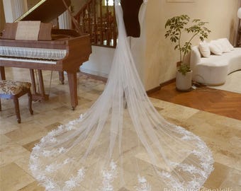 May include: A white bridal veil with a long train and floral lace trim. The veil is draped over a mannequin and is displayed on a wooden floor.