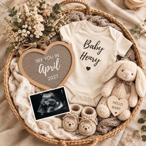 Puede incluir: Una cesta de mimbre que contiene un body de bebé color crema con el texto Baby Henry, un tablero de letras en forma de corazón que dice SEE YOU IN April 2027, un conejo de peluche beige con una etiqueta HELLO BABY, patucos de oso y una ecografía.