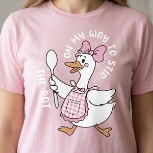 May include: Pale pink t-shirt with a cartoon goose design. The goose wears a pink bow and apron, holding a spoon. Text reads "No on my way to stir the pot". The design is centered on the shirt.