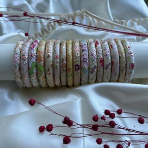 May include: A collection of floral-decorated bangles in various colors, including pink, purple, and green, are displayed. The bangles are arranged on a white cylindrical object against a backdrop of white fabric. Red berries and stems are in the foreground.
