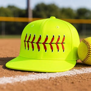 May include: A neon yellow baseball cap with a red and black stitched design, next to a yellow softball. The cap and ball are on a brown surface with a white line, suggesting a baseball field.